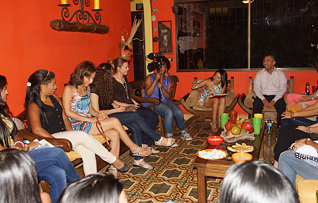 A television crew films a group of Colombian women seated in a cozy orange-walled living room during a romance tour interview session