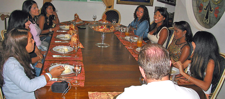 Dinner Introduction with Colombian Women A man sits at the head of a long wooden table surrounded by Colombian women during a formal matchmaking dinner meeting with wine glasses and conversation