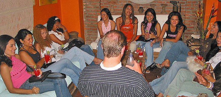 Casual Cocktail Meeting in Colombia A man enjoys drinks and laughter with a large group of Colombian women in casual clothes sitting around a low table in a cozy lounge with orange walls