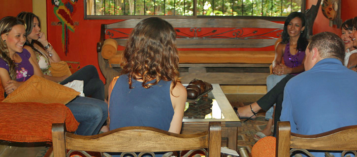 Small Group Matchmaking Conversation Several Colombian women and an American man seated together in a relaxed indoor setting with bright orange walls, smiling and enjoying casual conversation
