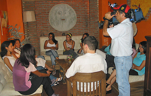 A Univision television crew films a lively discussion between Colombian women and an American man inside a warmly lit lounge during a matchmaking event