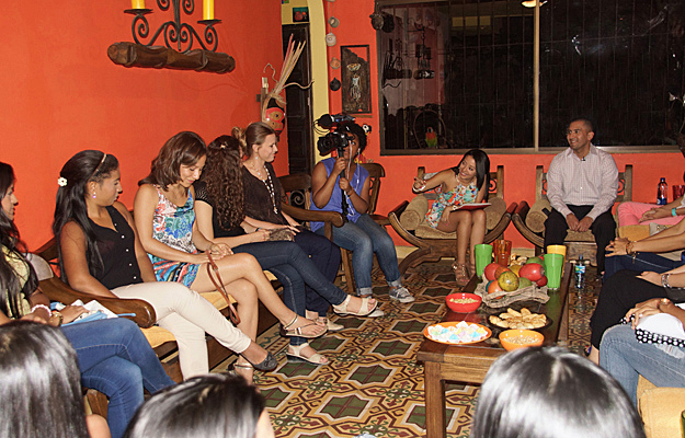 A television crew films a group of Colombian women seated in a cozy orange-walled living room during a romance tour interview session