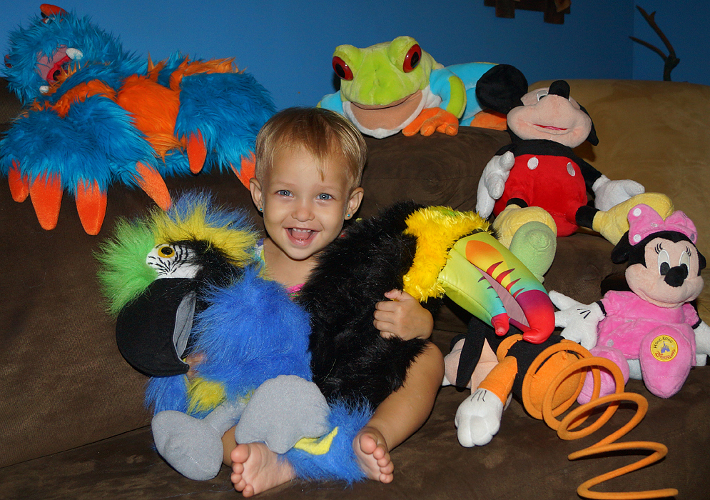 Beautiful Colombian American toddler sitting and smiling on a couch filled with colorful stuffed animals