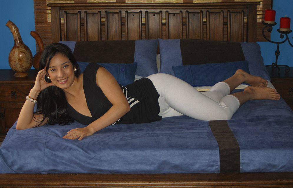Blakc hair, light skin Latin woman wearing white pants and a black shirt lying on a bed with blue and brown covers 