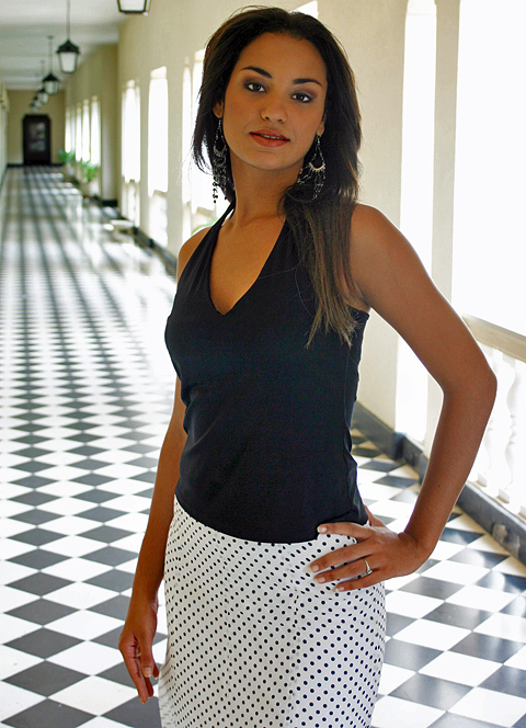 Smiling Colombian woman with long black hair wearing a fitted black tank top and blue jeans, standing in front of a wooden cabinet and artwork in a warmly lit room