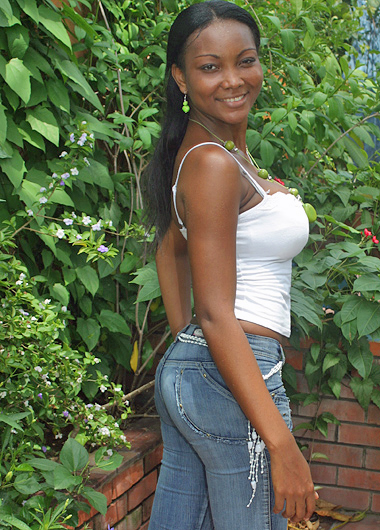 Colombian woman with long black hair wearing a white tank top and blue jeans, smiling and posing sideways near thick green plants