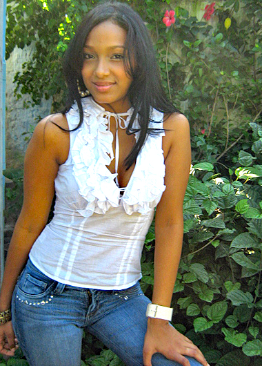 Colombian woman with long black hair wearing a white ruffled sleeveless top and blue jeans, posing outdoors beside lush green foliage