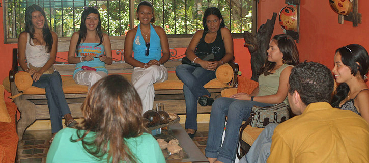 Group of young Colombian women sitting on a sofa and talking with a man during an introduction event inside a warm, orange-colored room with tropical decor
