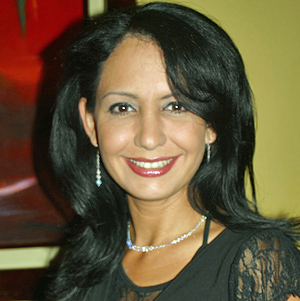 A mature Colombian woman with long black hair, wearing a black lace blouse and silver jewelry, smiling indoors under warm lighting