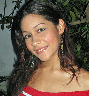 Latina Woman in Red Off-Shoulder Top Young Latina woman with light brown hair and dangling earrings, smiling softly while wearing a red off-shoulder top in an outdoor setting with plants behind her
