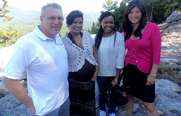 A Colombian woman stands smiling with her American husband, a pregnant belly visible, alongside two women from a television crew on a sunny hillside during an interview