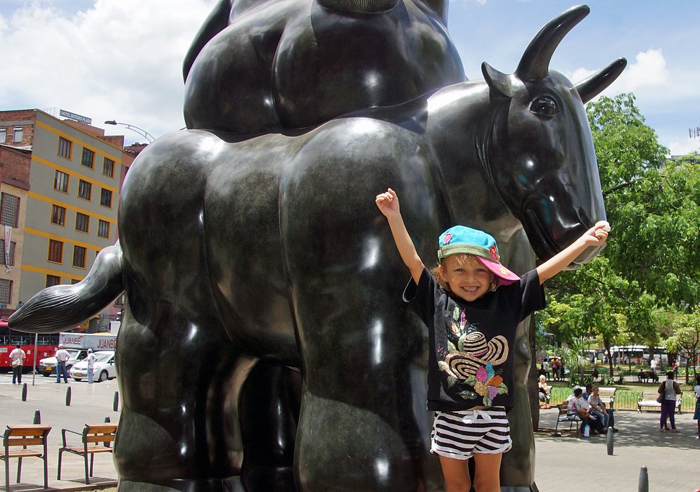 An attractive three year old holding her arms up in front of statue of a bull