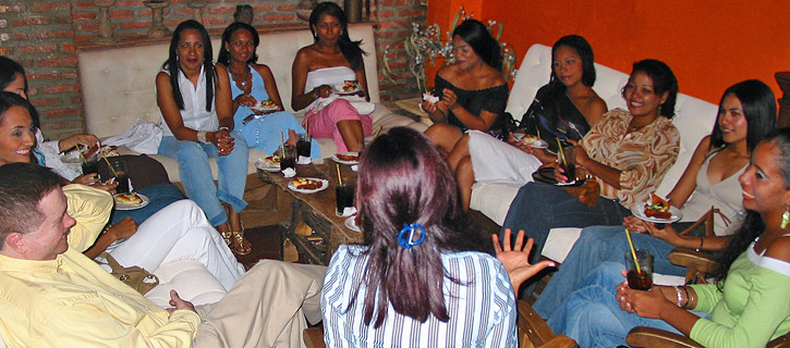Colombian Women Social Gathering A circle of Colombian women seated on sofas and chairs during an informal social event, holding drinks and small plates while listening to a man and engaging in relaxed conversation