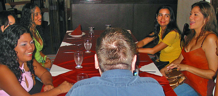 Colombian Dating Meeting Event A group of Colombian women gathered around a restaurant table with red tablecloths, smiling and talking while seated across from a man participating in a meet-and-greet