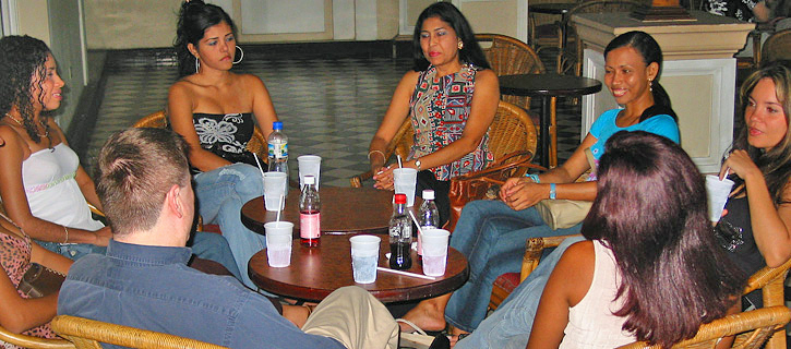 Colombian Women Personal Introductions A small group of Colombian women seated around a round coffee table with drinks in plastic cups, talking casually with a man during an informal introduction event