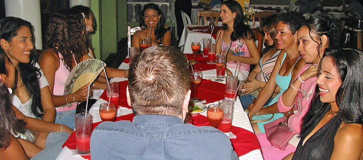 Colombian Women Dinner Introduction Event A lively group of Colombian women seated around a restaurant table with pink drinks and colorful outfits, smiling and chatting during a dinner introduction meeting with a visiting man