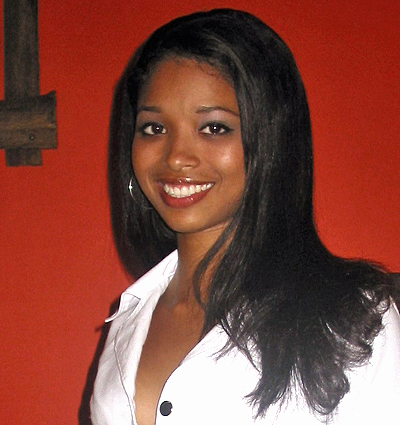 Smiling tan Latin woman with straight black hair wearing a white buttoned shirt, standing against an orange wall background