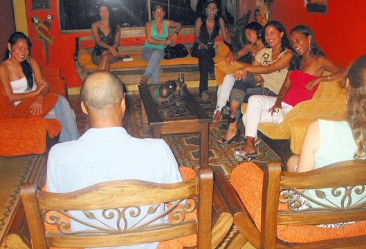 A Black man talks with smiling Colombian women seated in a living room with orange walls and tile floor during a matchmaking introduction