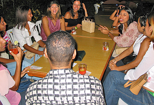 A group of Colombian women laugh and talk with a Black man at an outdoor table while enjoying drinks during a matchmaking gathering