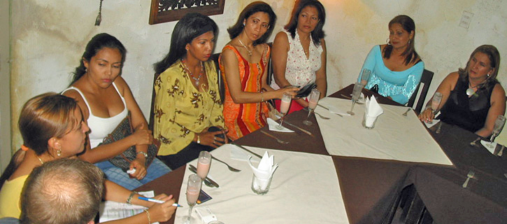 A group of women sitting at a restaurant table smiling and talking with a man during a social introduction event