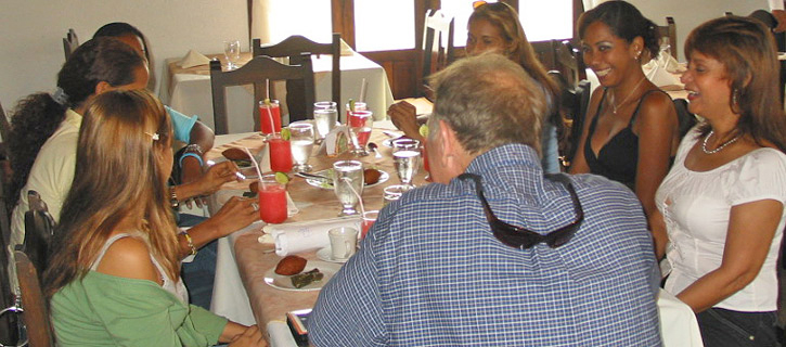 Several women seated around a round restaurant table listening to a man speak during a matchmaking introduction meeting