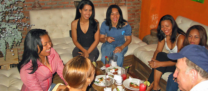 A group of women sitting at a long dining table listening to a man during a matchmaking meeting