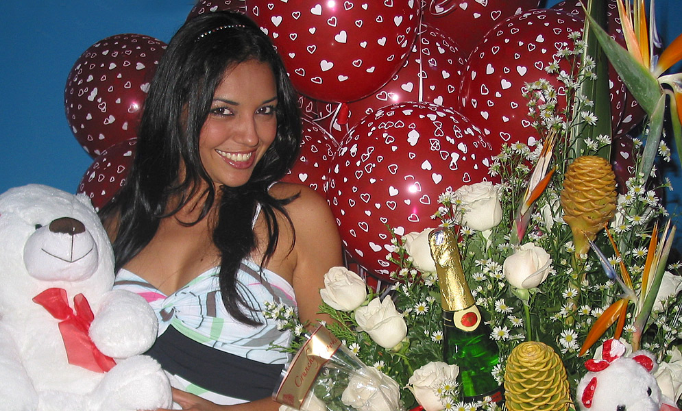 Dark hair, sexy Latin woman holding a white teddy bear and white roses with red ballons in the background