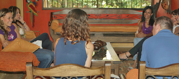 Several Colombian women and an American man seated together in a relaxed indoor setting with bright orange walls, smiling and enjoying casual conversation