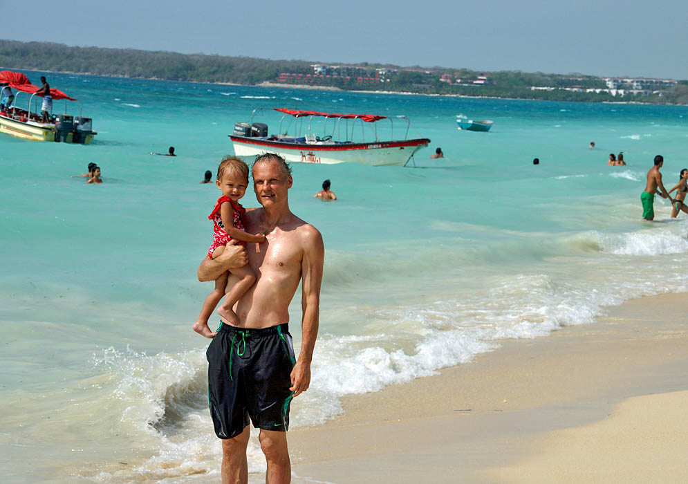 Father holding his two year old daughter on the seashore of Playa Blanca with blue water and boats in the background