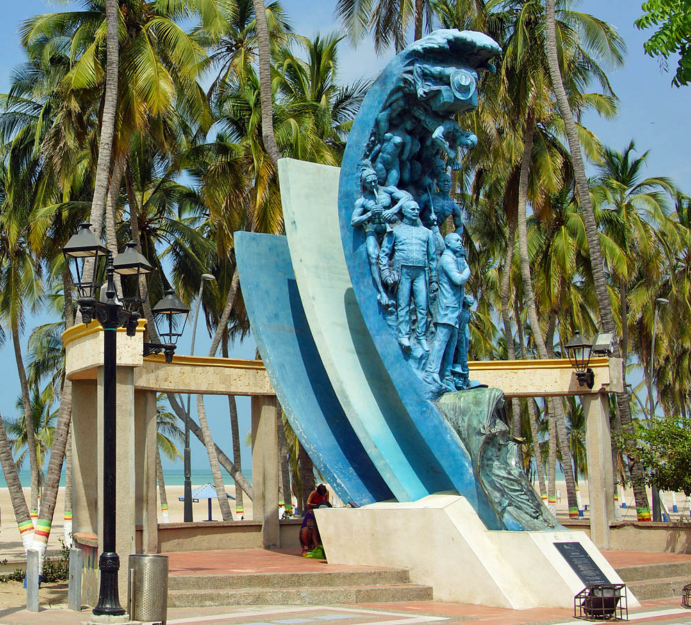 A blue statue representing historical figures of La Guajira with beach, ocean and palm trees in the background