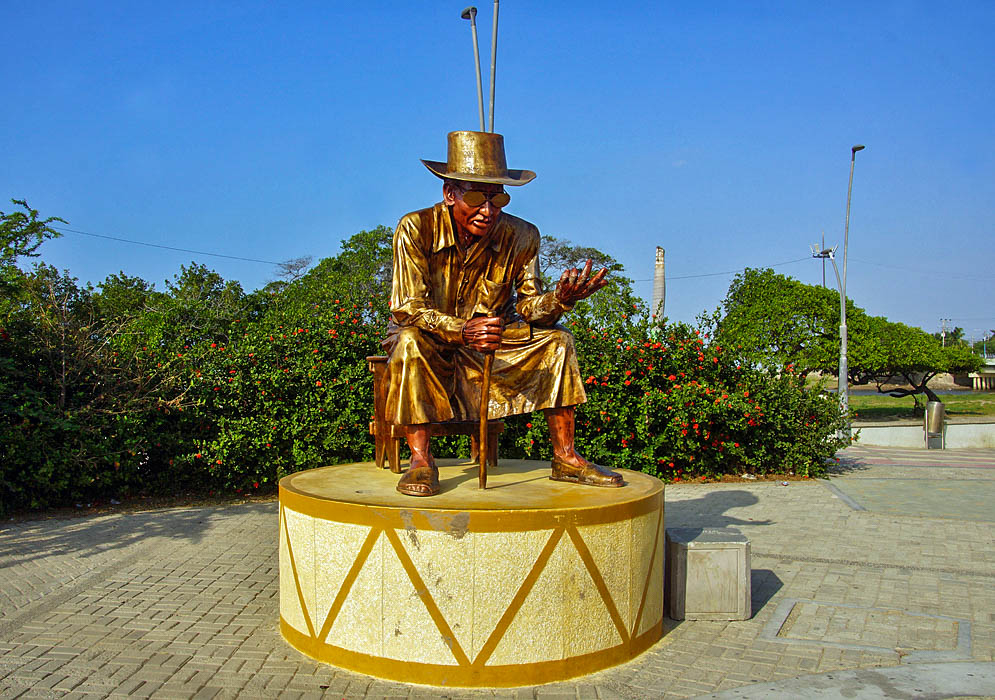A golden statue of a seated man with a hat, cane and glasses under blue skies
