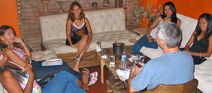A small circle of Colombian women chat with one guest in a cozy lounge with brick walls during a private introductions session