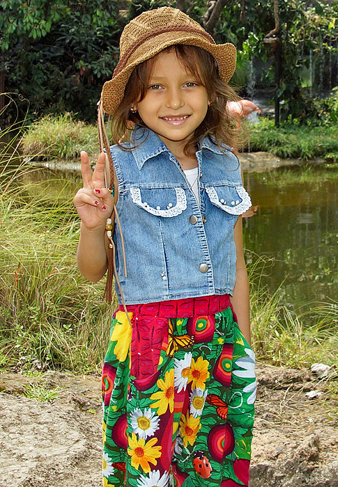 Stylish little girl wearing a jean vest, a brown hat and colorful pants give the peace sign