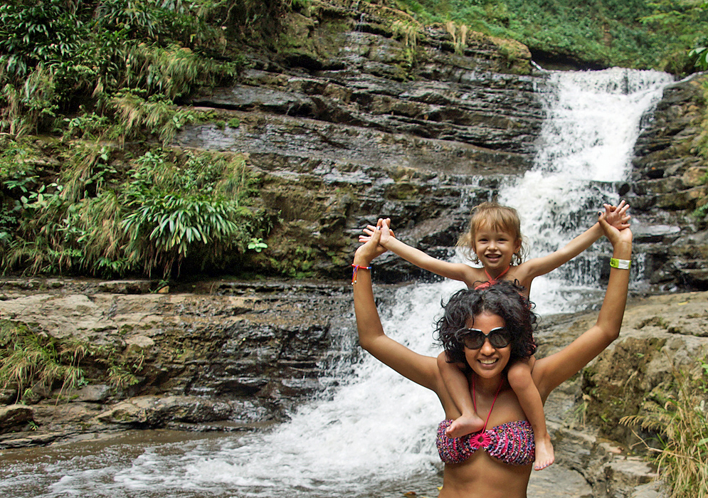 Three year old daughter on top of Colombian mom's shoulders in front of waterfall